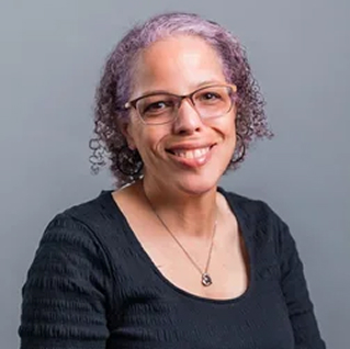 A portrait photo of Kat Shaw, a smiling black woman with silver hair with purple highlights, wearing glasses and a gold necklace.