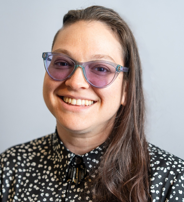 Smiling portrait of me, Marissa Epstein, a white woman with long brown hair, purple tinted glasses, a lack necklace, and a patterned shirt.